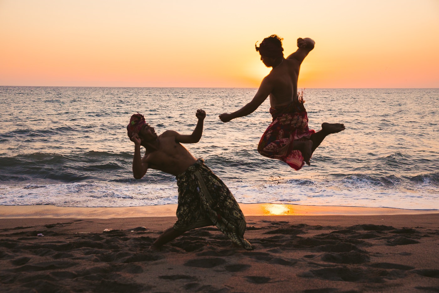 Two Sasak men in sarongs and headscarves performing a silat martial arts demonstration on a Lombok beach at sunset